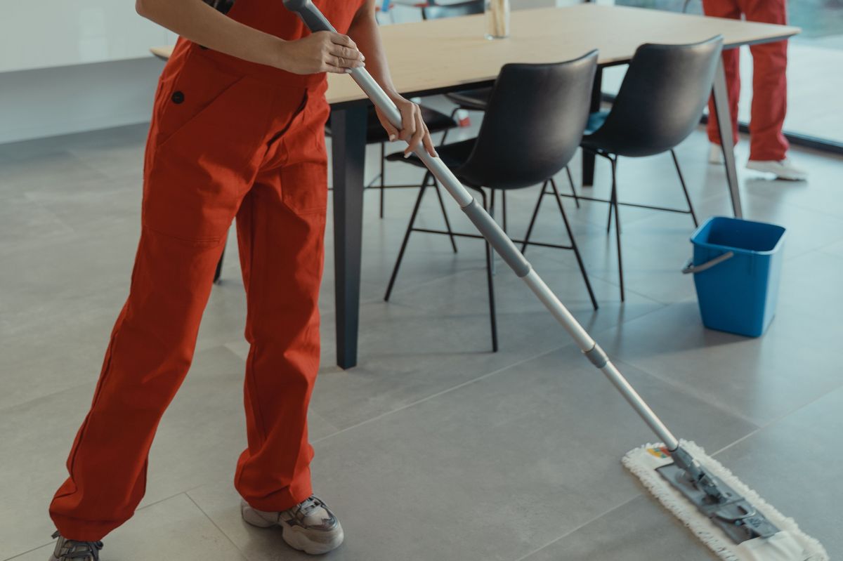 Iemand in een oranje werkoverall dweilt een grijze tegelvloer naast een eettafel