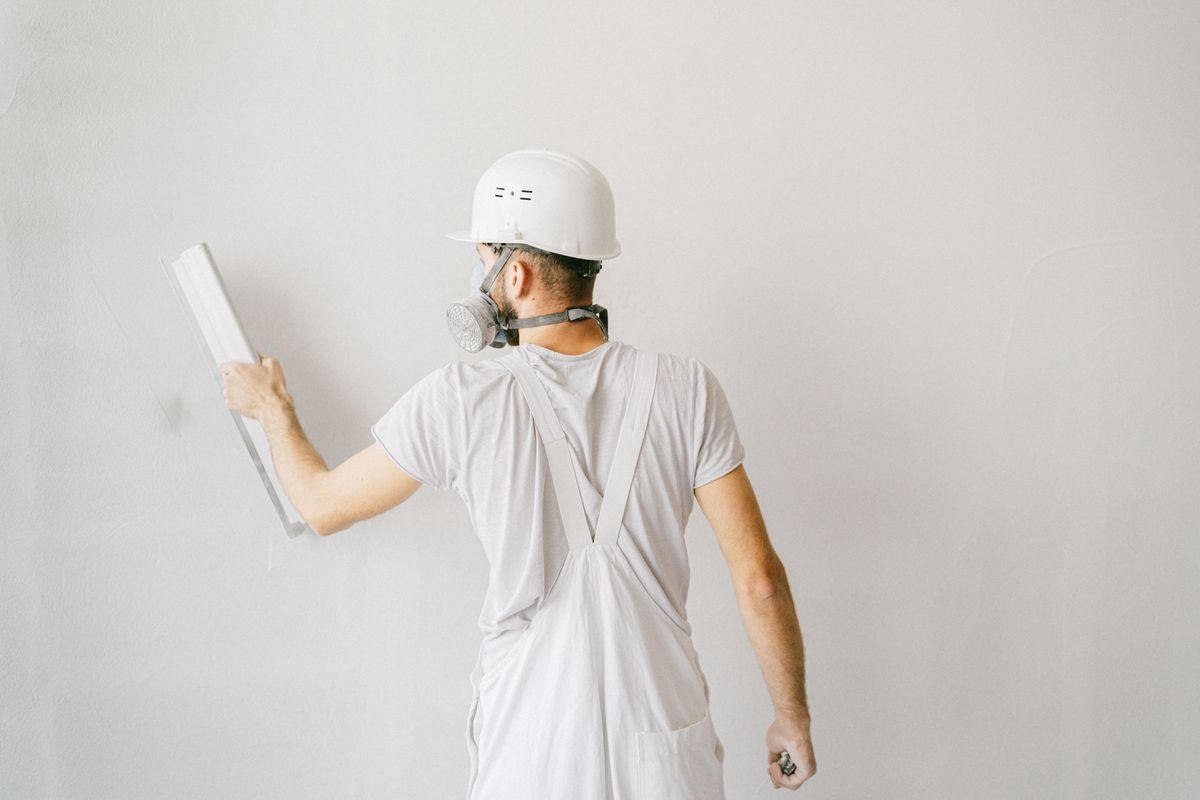 Stukadoor met helm en stofmasker strijkt een witte muur glad met een spaan